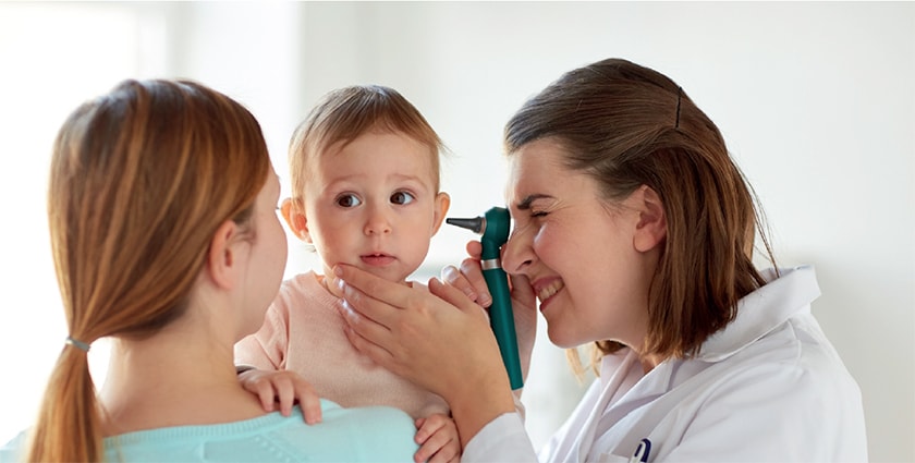 A mother holds her baby while a doctor checks its ears with an otoscope