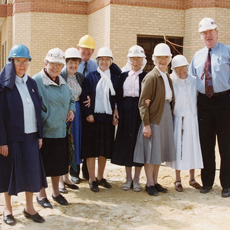 Sisters of St John of God wearing hard hats and standing on sand in front of the Murdoch hospital under construction