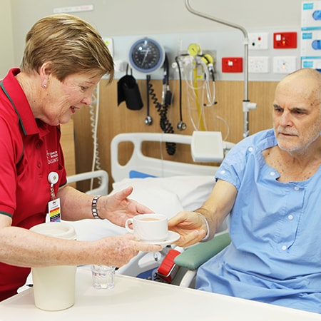 Volunteer wearing a uniform and badge passes a cup of tea to a patient wearing gown in bed
