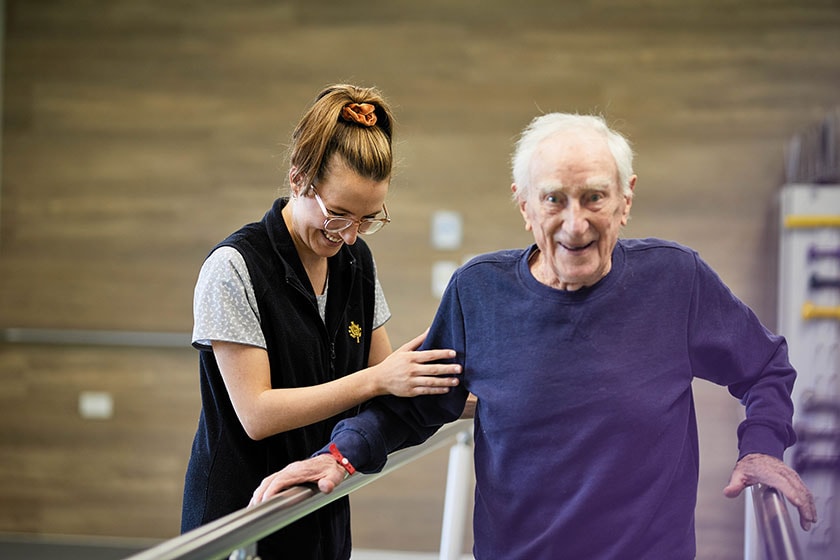 Caregiver assisting an elderly patient by holding their arm whilst they walk using the parallel bars for assistance in a rehabilitation room.