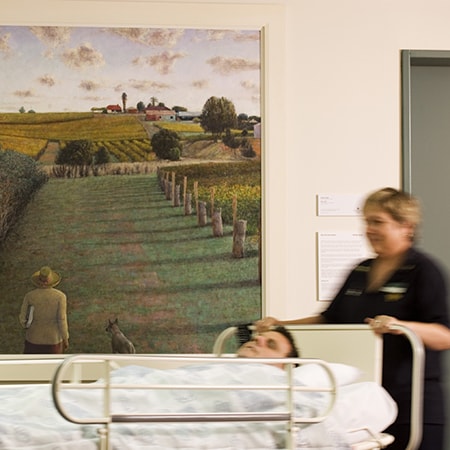 A work from the Murdoch Art Collection hanging on wall in hospital corridor as two caregivers wheel a patient in bed past it